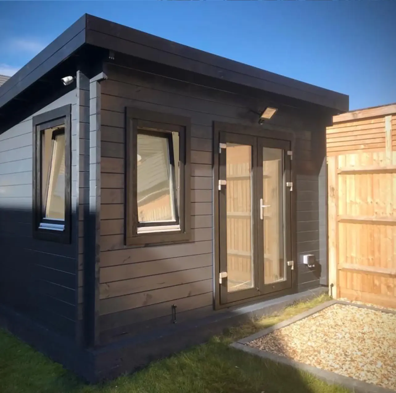 Compact black garden office with two tilt windows and double glass doors next to a wooden fence.